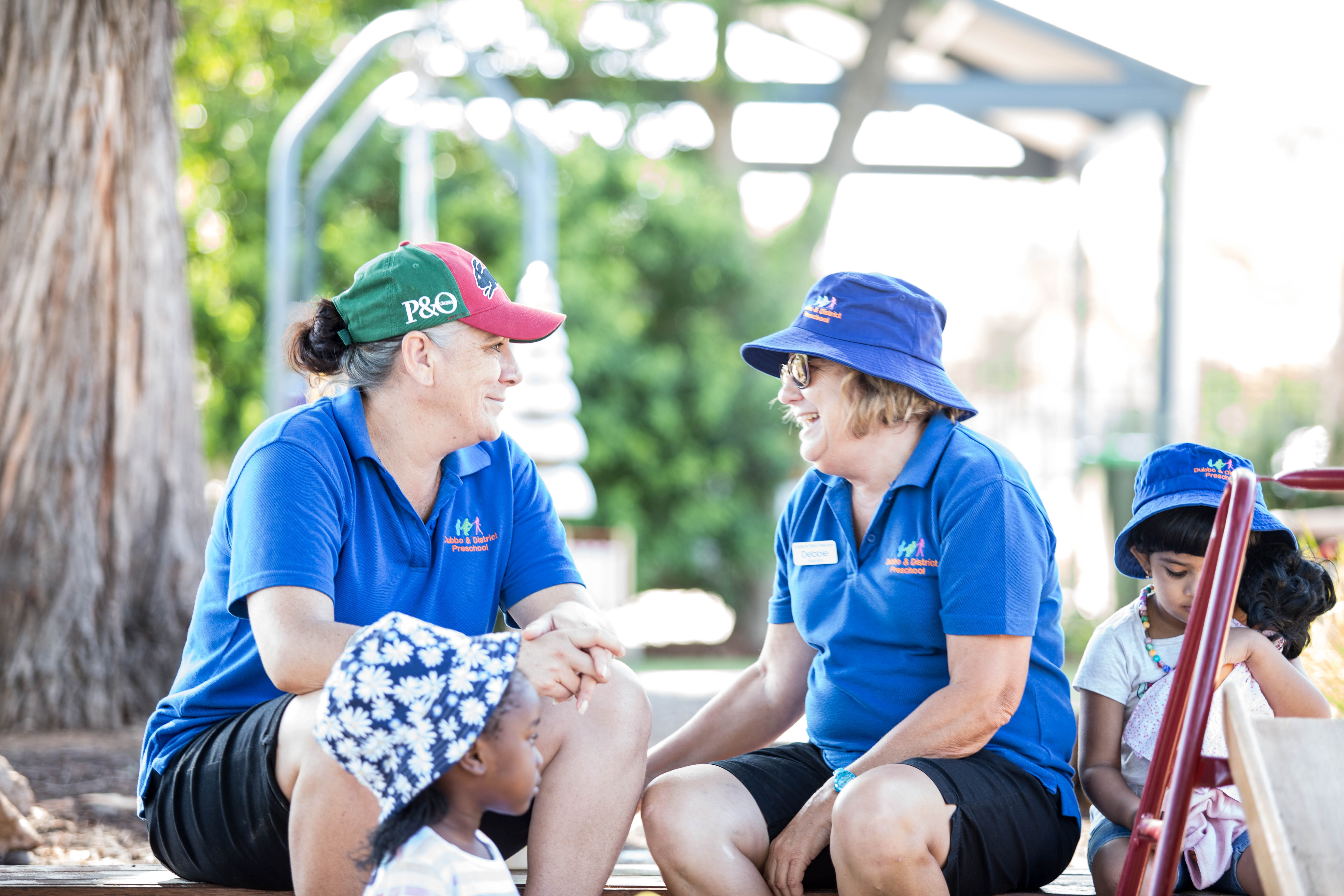 Two educators speaking in a playground, with young children 